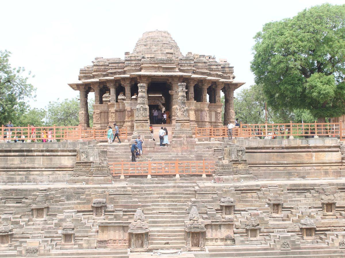Intricately carved ancient stone temple with a stepped base at the Dehra Mandir site, surrounded by trees and visitors