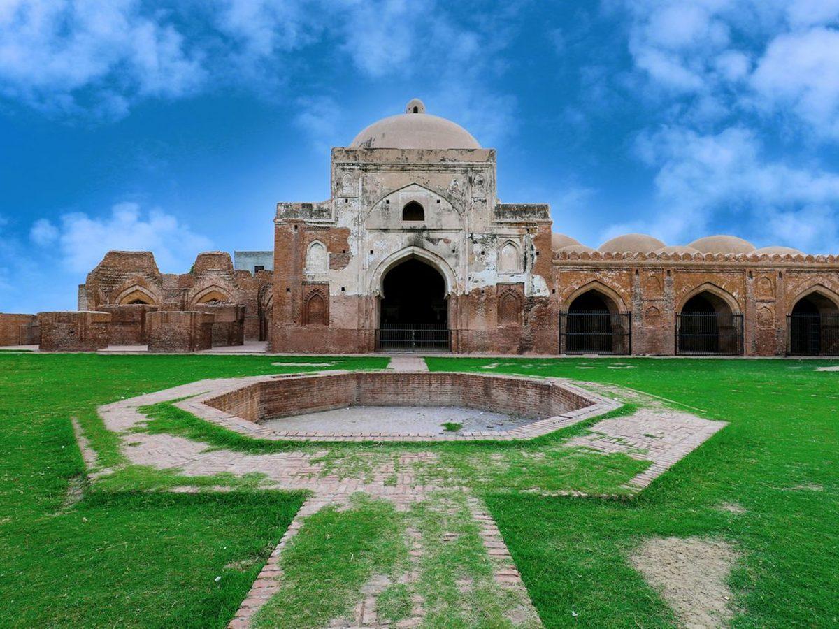 Ancient domed brick structure with arched openings at the Sheesh Mahal site, set against a clear blue sky with a green lawn in the foreground